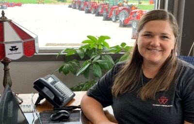 Elizabeth at her desk at Lauf Equipment, a row of tractors on the lot through the window behind her.
